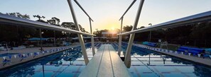 Una piscina con carriles, una plataforma de clavados y el sol poniéndose al fondo.