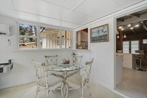 A dining area with a glass table surrounded by wicker chairs, a view of the outdoors through large windows, and a kitchen visible through an open door. - Cozy 3BR Sarasota Bungalow near Lido & Siesta (Sarasota)