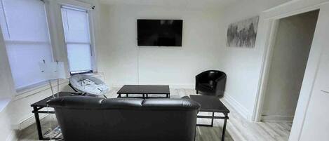 A modern living room with a black leather sofa, a flat-screen TV mounted on the wall, and a window with blinds.