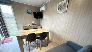 A room with a wooden dining table, black chairs, a blue sofa, a TV mounted on the wall, and a sliding glass door leading to an outdoor area. - The Ribouillet Cabin (CREON D'ARMAGNAC)