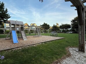 A playground with a slide and climbing structures, surrounded by a grassy area and a wooden fence. - Panoramic Ocean Views + Hot Tub: Steps to Beach (Lincoln City)