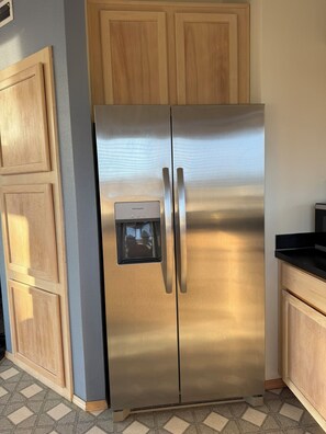 A stainless steel refrigerator with a water dispenser, situated in a kitchen with wooden cabinets and a tiled floor. - Panoramic Ocean Views + Hot Tub: Steps to Beach (Lincoln City)