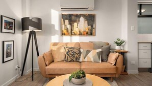 Una sala de estar moderna con sofá, lámpara de pie, un cuadro y una zona de cocina.