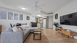 A modern living room with a sofa, a coffee table, a TV, and a kitchen area in the background. - Breathtaking! 2bed/2bath/1car/balcony/views ~ CBD (Brisbane)