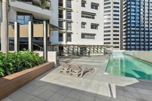 Ein Poolbereich auf dem Dach mit Liegestühlen, einem Holz-Pflanzgefäß mit Grünpflanzen und einem Hochhaus im Hintergrund.