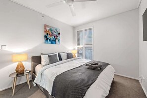 Ein Schlafzimmer mit einem Bett, einem Nachttisch, einer Lampe, einem Deckenventilator, einem Fenster mit Jalousien und einem Bild an der Wand.