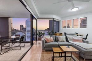 Ein modernes Wohnzimmer mit einer Couch, einem Couchtisch und Sesseln, ein Balkon mit Blick auf die Stadt und ein Deckenventilator.