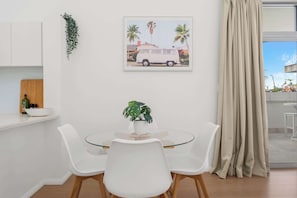 Ein Essbereich mit einem Glastisch, weißen Stühlen, einer Topfpflanze und einem gerahmten Bild an der Wand.