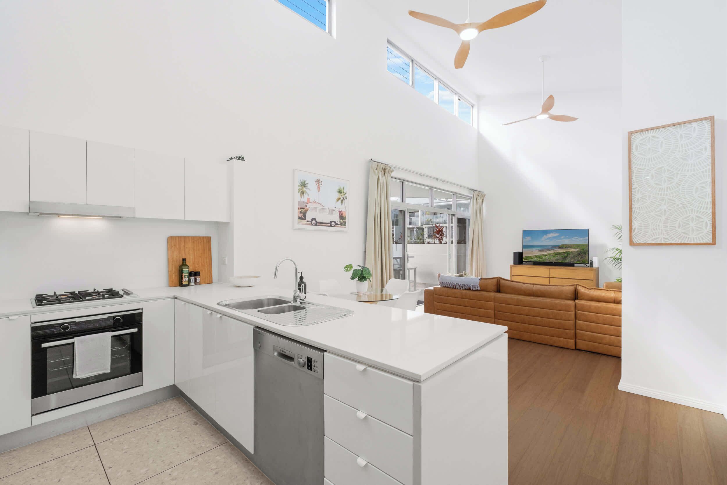 Una cocina moderna con armarios blancos, lavavajillas y estufa. Hay una zona de comedor con un sofá marrón y una televisión.