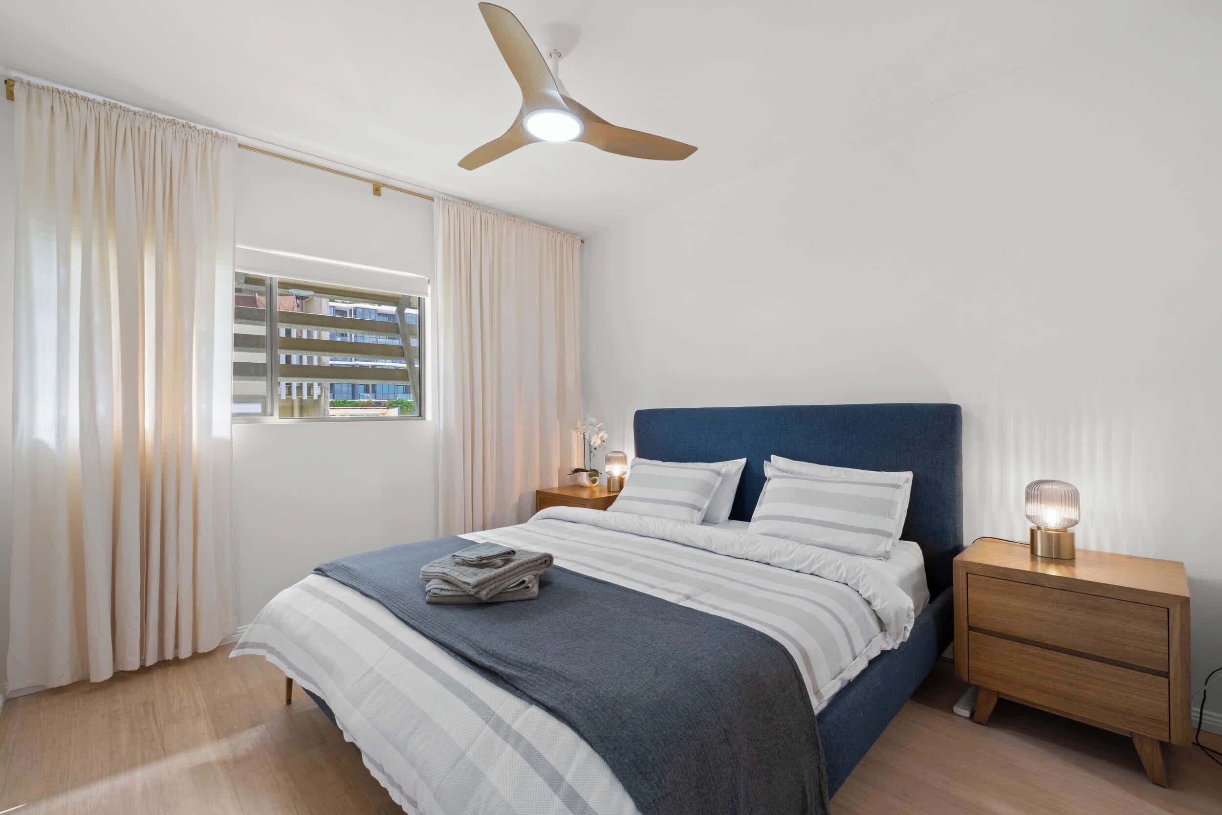 A bedroom with a bed, a nightstand, a ceiling fan, and a window with curtains.