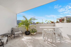 Un balcón con una mesa y sillas blancas, una planta en maceta y vistas de la ciudad.