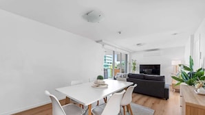 Uma sala de estar moderna com uma mesa de jantar branca, cadeiras brancas, um sofá escuro e uma televisão.