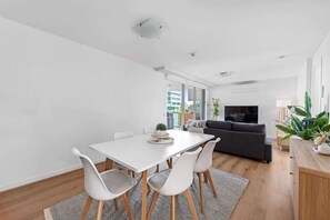 A modern living room with a white dining table, white chairs, a dark sofa, and a TV. - Inner City Action! 3bed/2bath/1car/3balconies (Brisbane)