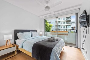 A modern bedroom with a bed, bedside tables, a ceiling fan, a TV, and a balcony with a view of buildings. - Inner City Action! 3bed/2bath/1car/3balconies (Brisbane)