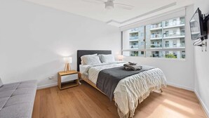 Um quarto com cama, mesa de cabeceira, abajur, ventilador de teto, janela com vista para outros edifícios e televisão fixada na parede.
