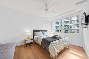 A bedroom with a bed, a nightstand, a lamp, a ceiling fan, a window with a view of other buildings, and a TV mounted on the wall. - Inner City Action! 3bed/2bath/1car/3balconies (Brisbane)