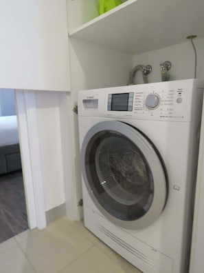 A front-loading washing machine in a room with a white door and tiled floor. - Central & Stylish! 1bed/1bath/1car ~ 5 Mins to CBD (Brisbane)