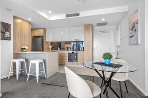 A modern kitchen with white cabinets, a central island, bar stools, and a glass dining table with chairs. - City Living! 1bed/1bath/pool (Brisbane)