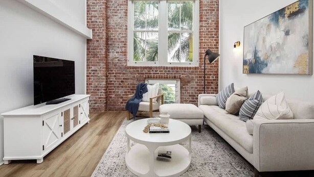 A modern living room with a brick wall, a large window, a flat-screen TV, a white sofa, and a coffee table. - Woolstore Style! ~ 2bed/2bath/1car ~ Teneriffe (Brisbane)