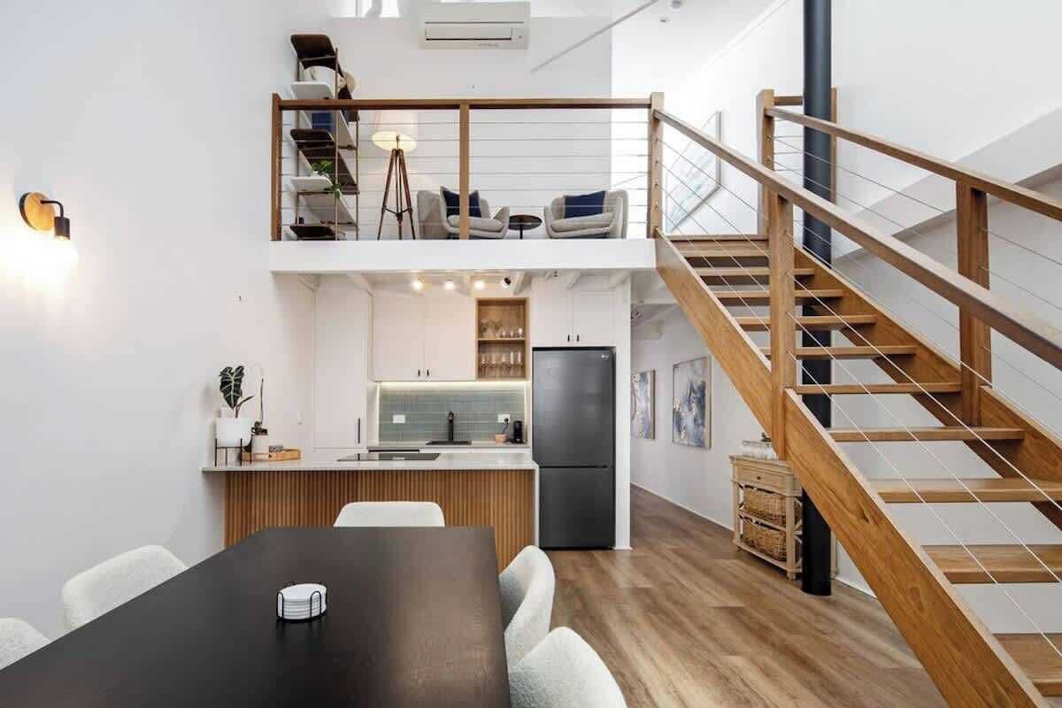Et moderne køkken med en central ø, et spiseområde med et sort bord, og en trappeopgang til et opholdsområde oppe.