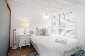 Ein Schlafzimmer mit Bett, Nachttisch, Lampe und einem Fenster mit Jalousien.