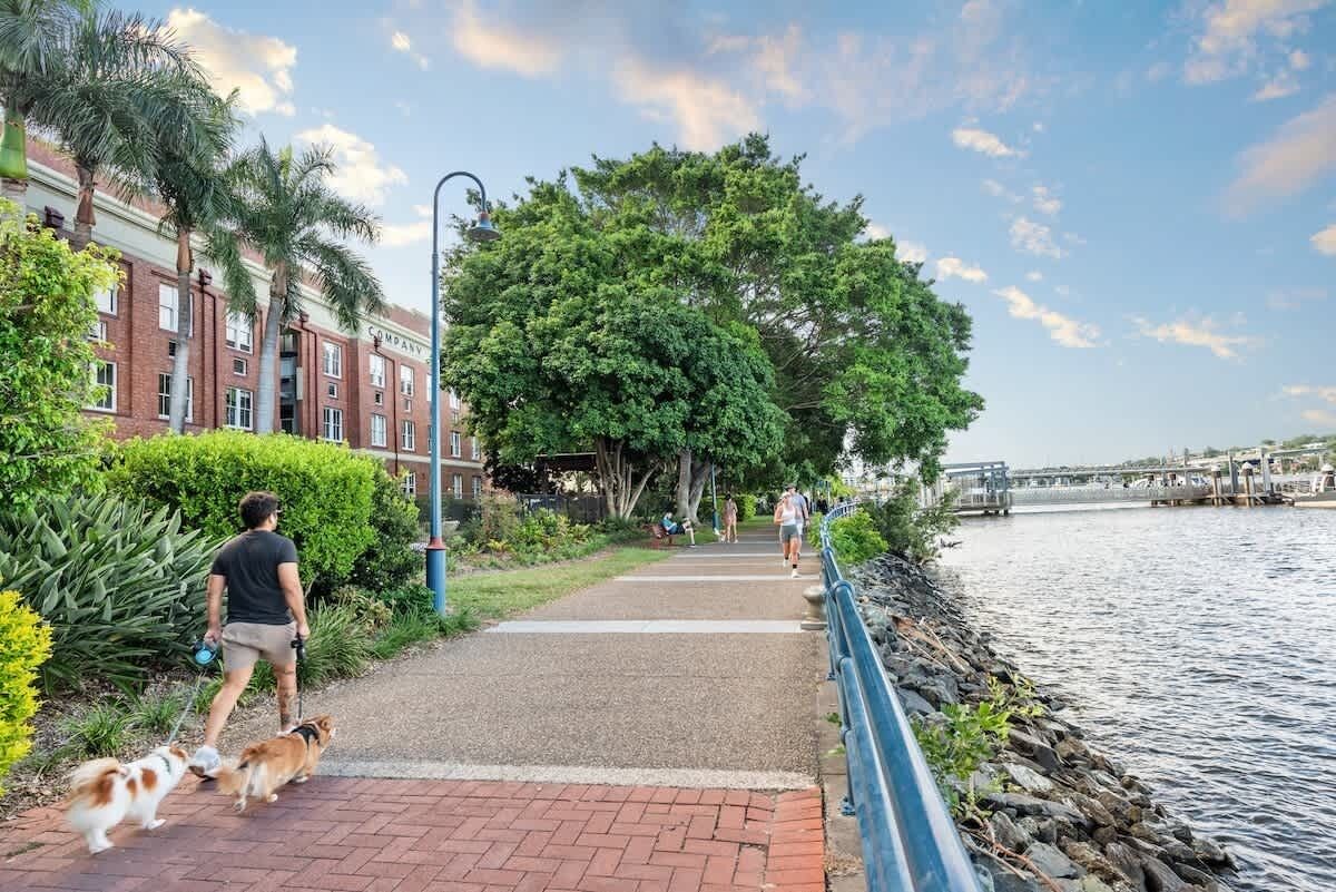 A pathway alongside a river with a brick walkway, trees, and a building with a sign.