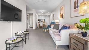A modern living room with a flat-screen TV, a ceiling fan, a sofa, a coffee table, a dining area, and a kitchen in the background. - Modern Style! 1bed/1bath/balcony/pool ~ CBD (Brisbane)