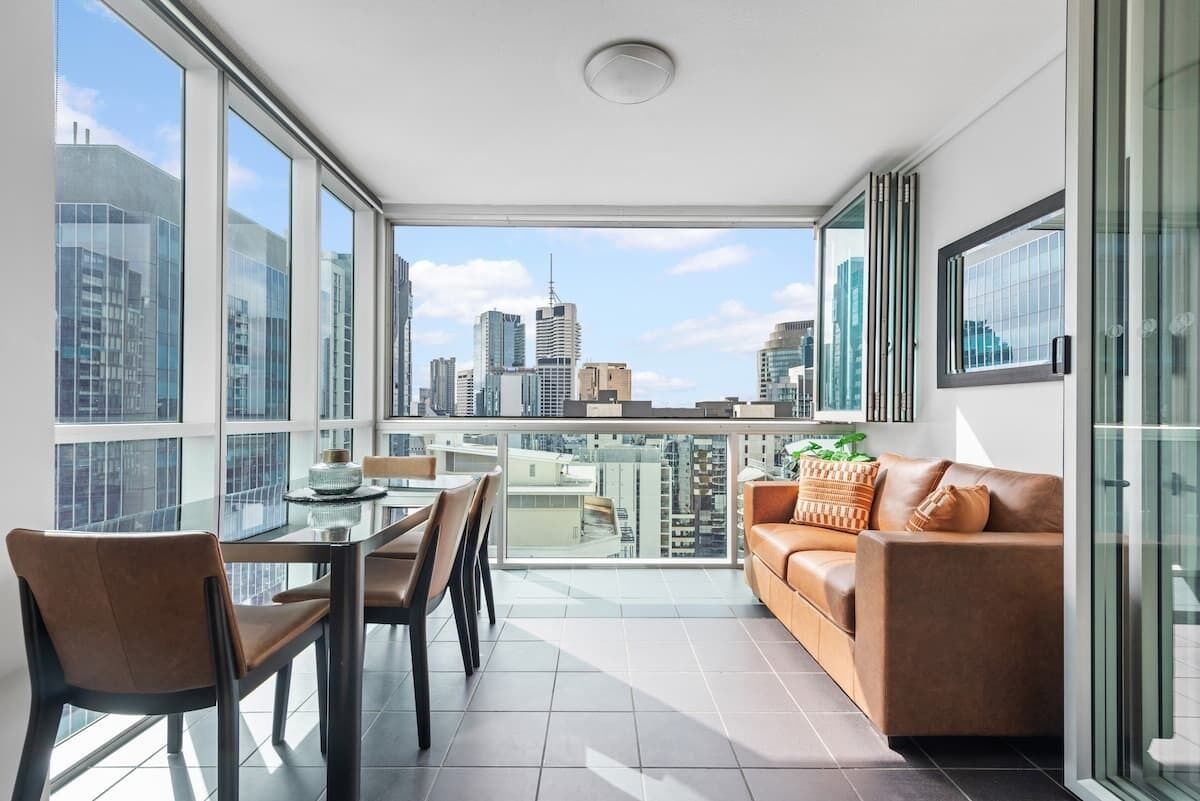 Una sala de estar moderna con una mesa de vidrio, sofá de cuero marrón y vistas a la ciudad a través de amplias ventanas.