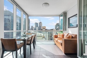 A modern living room with a glass table, brown leather sofa, and a city view through large windows. - Hot in the City! 2bed/2bath/2car/study/pool/views (Brisbane)