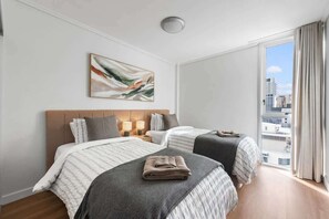 Ein Hotelzimmer mit zwei Betten, einem großen Fenster mit Blick auf Gebäude, einer Deckenleuchte und einem Gemälde an der Wand.