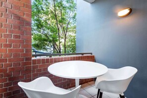 Eine Terrasse mit rundem Tisch und zwei weißen Stühlen, eine Ziegelwand und Blick auf Bäume durch ein Fenster.