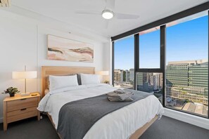 Ein modernes Hotelzimmer mit einem großen Bett, einem Nachttisch mit Lampe, einem Deckenventilator und Blick auf die Stadt durch große Fenster.