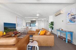 A modern living room with a sofa, coffee table, and a small stool. There is a dining area with a table and chairs, and a kitchen in the background. - Immaculate Top Floor Views! 2bed/2bath/1car/pool (Brisbane)