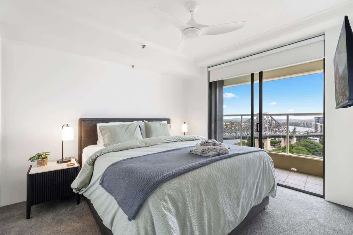 A modern hotel room with a large bed, a nightstand with a lamp, a ceiling fan, and a view of a cityscape through large windows.