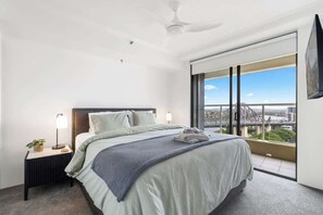 Ett modernt hotellrum med en stor säng, ett nattduksbord med en lampa, en takfläkt och utsikt över stadsbilden genom stora fönster.