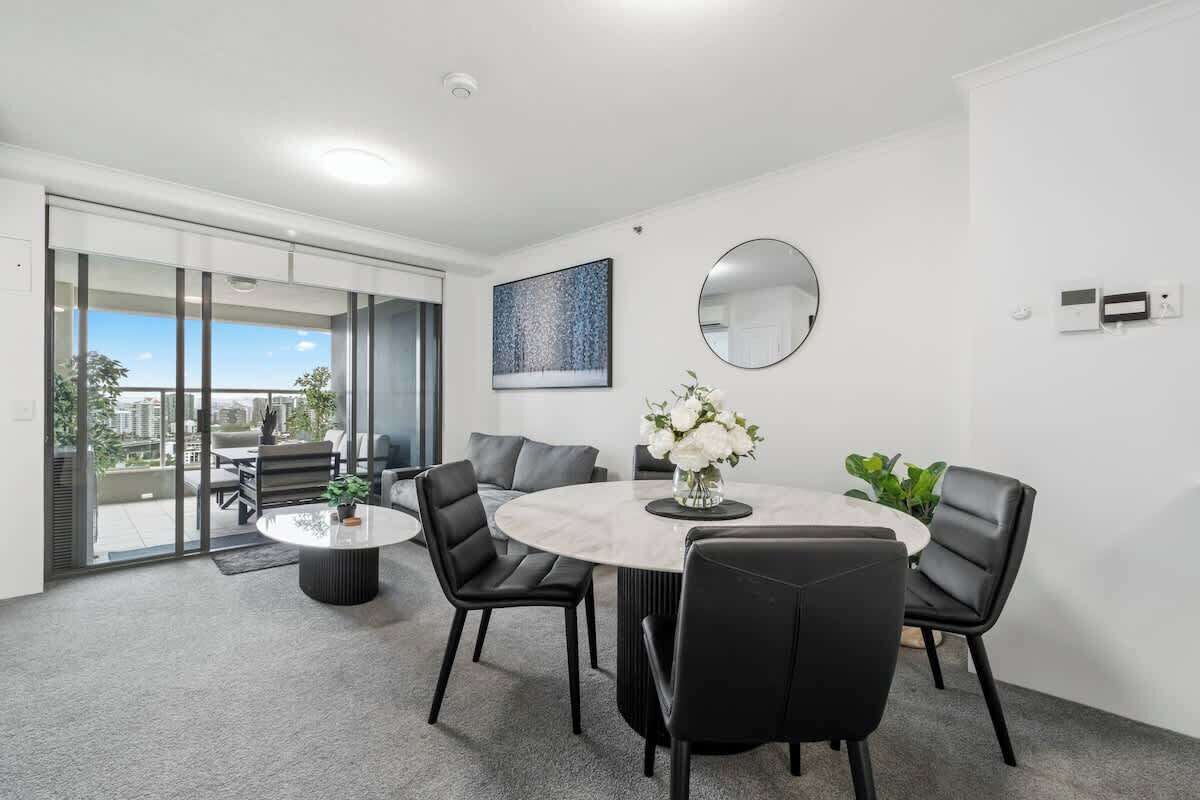Un espace de repas moderne avec une table ronde, des chaises noires, un vase de fleurs, un miroir rond et une vue sur la ville à travers de grandes fenêtres.