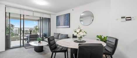A modern dining area with a round table, black chairs, a vase of flowers, a round mirror, and a city view through large windows.