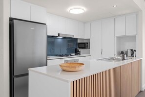 A modern kitchen with white cabinetry, a stainless steel refrigerator, a wooden bowl on the counter, and a built-in microwave. - City Escape! 2bed-2bath-1car-pool-views - CBD (Brisbane)