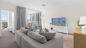 A modern living room with a grey sofa, a dining area with chairs, a large painting, and a view through a glass door. - City Escape! 2bed-2bath-1car-pool-views - CBD (Brisbane)