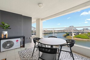 Ein Balkon mit Tisch und Stühlen, einem Ventilator, einem Blumentopf und Blick auf eine Brücke und einen Fluss.