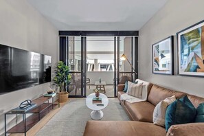 Ein modernes Wohnzimmer mit Flachbildfernseher, einer Couch, einem Couchtisch und einem Essbereich, der durch Glastüren sichtbar ist.