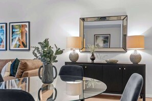 Ein modernes Wohnzimmer mit einem Glastisch, schwarzen Stühlen, einem braunen Sofa mit Kissen, einem dunklen Holzschrank und zwei Wandleuchten.