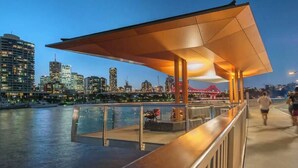 Eine Uferpromenade mit überdachten Sitzbereichen, Blick auf den Stadtpanoramahimmel und einer Brücke im Hintergrund.
