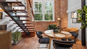 Ein modernes Interieur mit einer Ziegelwand, einer Holztreppe, einem runden Esstisch mit Stühlen und einem großen Fenster.