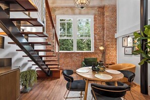 A modern interior with a brick wall, a wooden staircase, a round dining table with chairs, and a large window. - Woolstore Elegance ~ 2bed/2bath/1car ~ Teneriffe (Brisbane)