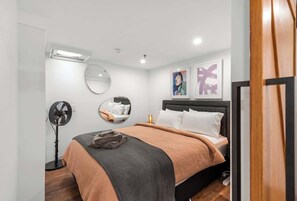A modern bedroom with a bed, bedside table, mirror, artwork, and a standing fan. - Woolstore Elegance ~ 2bed/2bath/1car ~ Teneriffe (Brisbane)