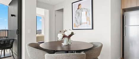 A dining area with a round table, four grey chairs, a vase with white flowers, and a framed picture on the wall.