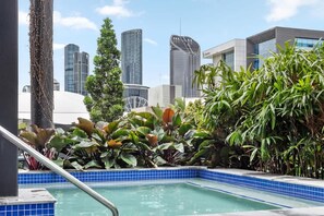 Ein Pool auf dem Dach mit blauen Fliesen, umgeben von tropischen Pflanzen und modernen Gebäuden im Hintergrund.