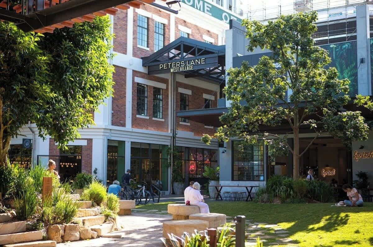 An outdoor seating area with stone planters, a covered seating area, and a building labeled 'Peters Lane West Village' with visible storefronts.