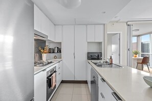A modern kitchen with white cabinetry, a built-in oven, a dishwasher, and a large island with a sink. - Top Floor! 2bed/2bath/1car/pool/views ~ CBD (Brisbane)